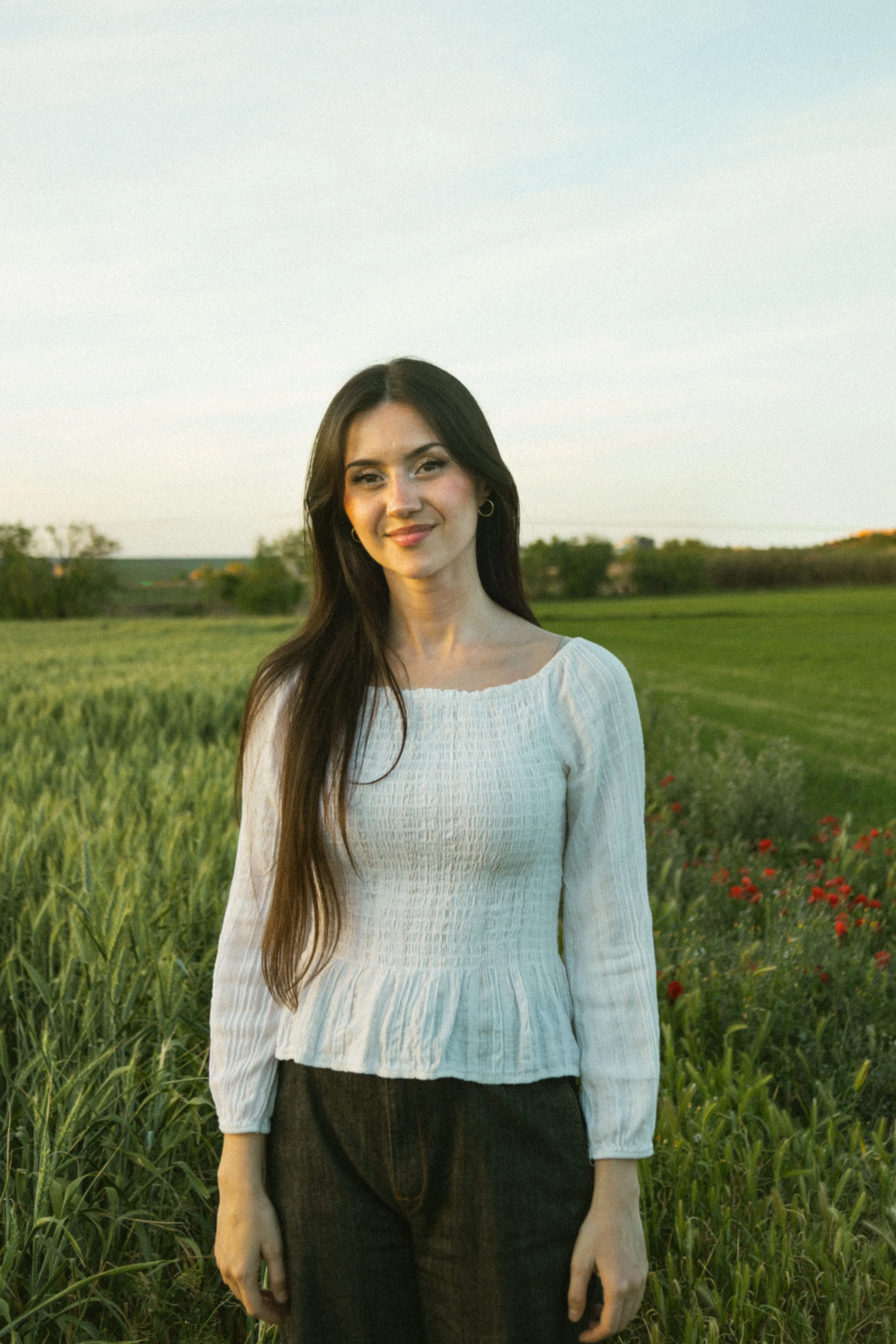 Paula Burriel sonriendo, blusa blanca, junto a un trigal con amapolas al atardecer.